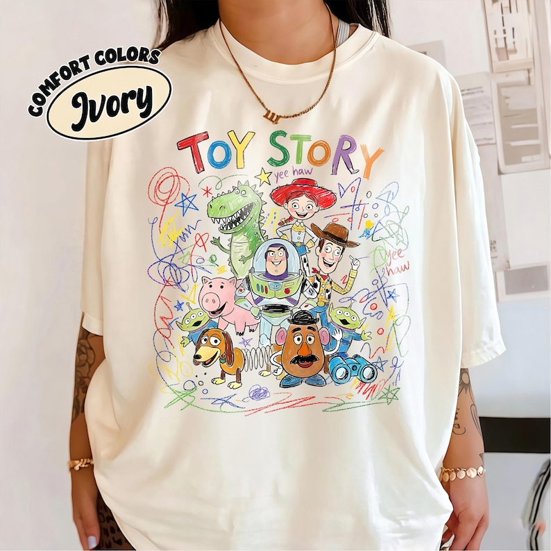 K&ouml;nnte beinhalten: Cremefarbenes T-Shirt mit einer farbenfrohen Grafik von Toy Story-Charakteren. Das Design zeigt Woody, Buzz Lightyear und andere Charaktere aus dem Animationsfilm, mit den Worten "TOY STORY" in einer verspielten Schriftart.