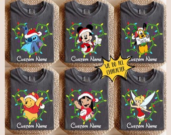 Puede incluir: Camisetas grises con varios personajes de dibujos animados con gorros de Papá Noel, rodeados de luces navideñas. Cada camiseta tiene el texto "Custom Name" debajo. Un banner amarillo en forma de estrella dice "WE DO ALL CHARACTER".