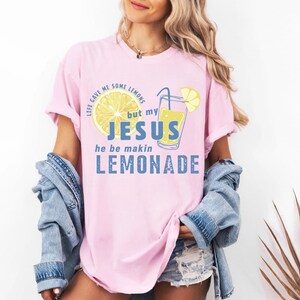 May include: A light pink t-shirt with the text "LIFE GAVE ME SOME LEMONS but my JESUS he be makin LEMONADE" in a mix of fonts and colors. The design includes a lemon slice and a glass of lemonade.