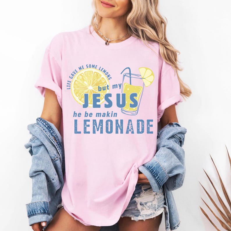 May include: A light pink t-shirt with the text "LIFE GAVE ME SOME LEMONS but my JESUS he be makin LEMONADE" in a mix of fonts and colors. The design includes a lemon slice and a glass of lemonade.
