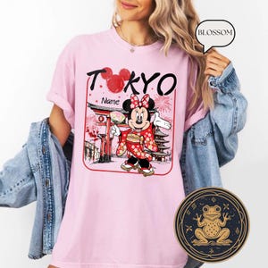 May include: Pink t-shirt with a graphic of Minnie Mouse in a kimono, holding a fan, in front of a Japanese temple and the word "TOKYO". The shirt also has a round frog design.