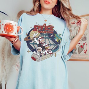 K&ouml;nnte beinhalten: Ein hellblaues T-Shirt mit einem Vintage-Grafikdruck von Mickey Mouse, Goofy und Pluto, die eine Space Mountain-Achterbahn fahren. Die Grafik enth&auml;lt den Text "SPACE MOUNTAIN" in roten und wei&szlig;en Buchstaben.