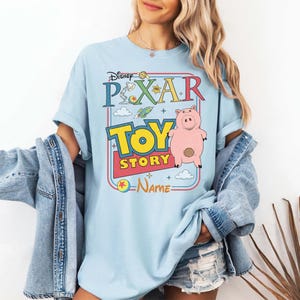 Peut inclure: T-shirt bleu clair avec le logo Disney Pixar Toy Story et un dessin de cochon rose. Le design comprend le texte "TOY STORY" en jaune et rouge, avec le logo Pixar en haut. Le t-shirt a des manches courtes.
