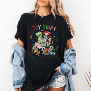 K&ouml;nnte beinhalten: Schwarzes T-Shirt mit einer farbenfrohen Grafik von Toy Story-Charakteren, darunter Woody und Buzz Lightyear. Der Text "TOY STORY" ist in Regenbogenfarben &uuml;ber den Figuren geschrieben. Das Design ist von bunten Kritzeleien umgeben.
