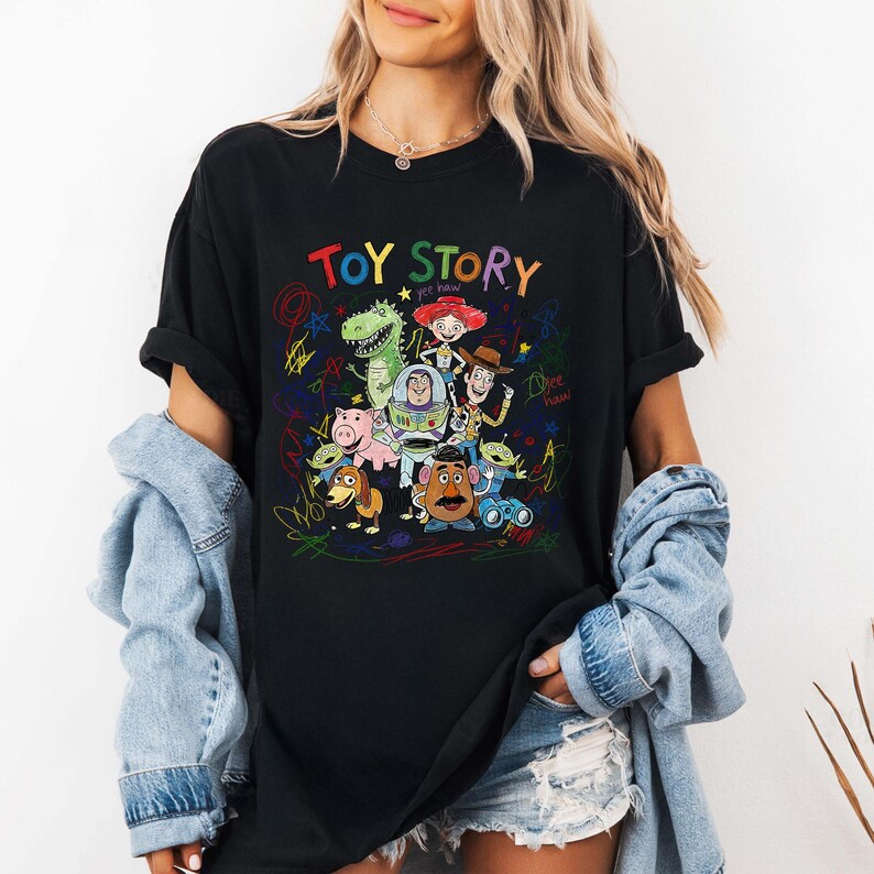 K&ouml;nnte beinhalten: Schwarzes T-Shirt mit einer farbenfrohen Grafik von Toy Story-Charakteren, darunter Woody und Buzz Lightyear. Der Text "TOY STORY" ist in Regenbogenfarben &uuml;ber den Figuren geschrieben. Das Design ist von bunten Kritzeleien umgeben.