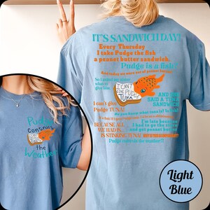 Puede incluir: Camisetas de color azul claro con un gr&aacute;fico de un pez naranja y el texto "Pudge Controls the Weather" y "I can't give Pudge Tuna!". La parte trasera de la camiseta tiene un dise&ntilde;o humor&iacute;stico con tem&aacute;tica de s&aacute;ndwich.