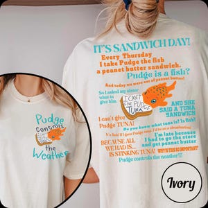 Puede incluir: Camiseta de color marfil con el texto "Pudge Controls the Weather" y una ilustraci&oacute;n de pez de dibujos animados. La parte trasera de la camiseta tiene un dise&ntilde;o humor&iacute;stico con tem&aacute;tica de s&aacute;ndwich con texto y un gr&aacute;fico de pez.