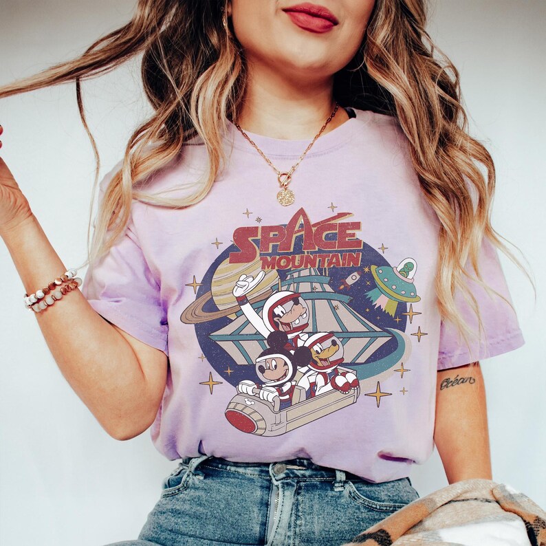 K&ouml;nnte beinhalten: Ein lilafarbenes T-Shirt im Tie-Dye-Look mit einer Vintage-Grafik von Mickey Mouse, Donald Duck und Goofy, die eine Space Mountain-Fahrt machen. Der Text "Space Mountain" ist auf dem Shirt aufgedruckt.