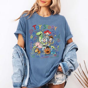 K&ouml;nnte beinhalten: Blaues T-Shirt mit dem Schriftzug "TOY STORY" in bunten Buchstaben. Das Shirt hat eine Grafik verschiedener Toy Story-Charaktere, darunter Woody, Buzz Lightyear und Rex, mit bunten Kritzeleien um sie herum.