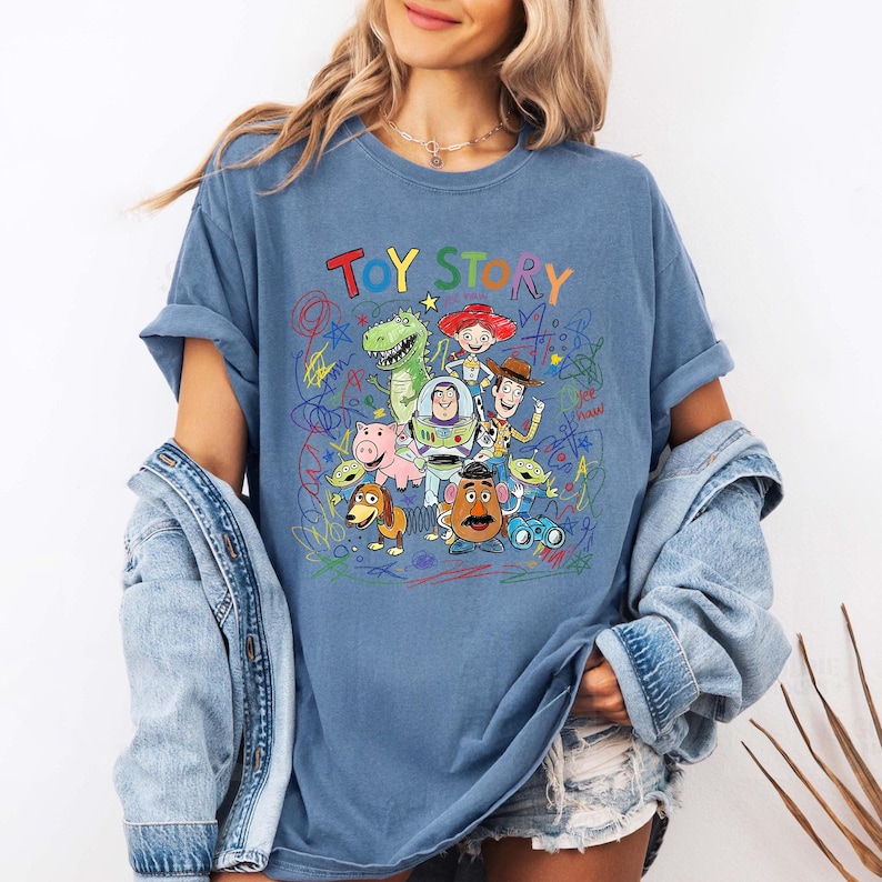 K&ouml;nnte beinhalten: Blaues T-Shirt mit dem Schriftzug "TOY STORY" in bunten Buchstaben. Das Shirt hat eine Grafik verschiedener Toy Story-Charaktere, darunter Woody, Buzz Lightyear und Rex, mit bunten Kritzeleien um sie herum.