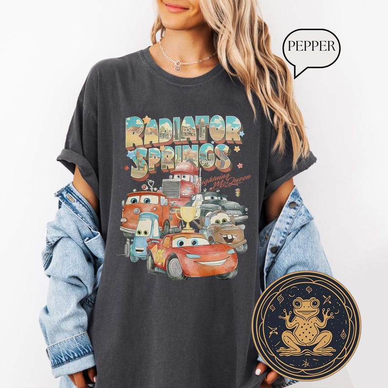 May include: A dark gray t-shirt with a vintage-style graphic of the Radiator Springs town from the Cars movie. The graphic features the text "Radiator Springs" and the characters Lightning McQueen and Mater.