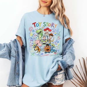 K&ouml;nnte beinhalten: Hellblaues T-Shirt mit dem Schriftzug "Toy Story" und einer farbenfrohen Grafik von Figuren aus dem Animationsfilm. Das Design umfasst Woody, Buzz Lightyear, Rex und andere beliebte Charaktere, umgeben von gekritzelten Linien.