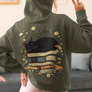 Puede incluir: Una sudadera con capucha verde con un gato negro durmiendo sobre una pila de libros. El gato está rodeado de flores amarillas y blancas.