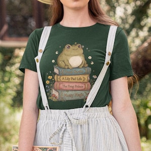 Könnte beinhalten: Dunkelgrünes T-Shirt mit einem freundlichen Frosch, der auf einem Bücherstapel sitzt, darunter "Frog Spells" und "A Lily Pad Life". Das Shirt hat ein verspieltes, illustriertes Design mit floralen Akzenten.