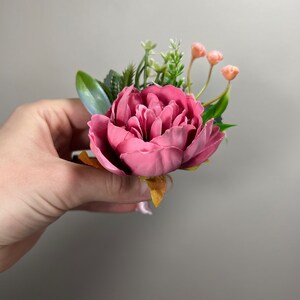May include: A pink peony flower with green leaves and small pink buds. The flower is arranged in a bouquet.