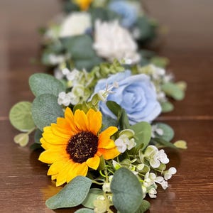 May include: A floral table runner featuring a bright yellow sunflower, light blue roses, white flowers, and green foliage. The arrangement is set on a dark wooden surface, creating a vibrant contrast.