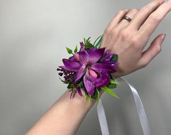 May include: A purple orchid wrist corsage with green leaves and a white satin ribbon.