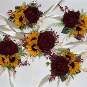 Könnte beinhalten: Fünf Ansteckblumen mit burgunderroter Rose und gelber Sonnenblume mit elfenbeinfarbenem Satinband.