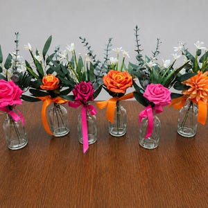Op de afbeelding: Zes kleine glazen vazen ​​houden elk een kleurrijk bloemstuk. De bloemen zijn in tinten roze, oranje en wit, met groen blad. Elke vaas is vastgebonden met een lint. De vazen ​​staan ​​op een bruin houten oppervlak.