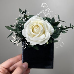May include: A single white rose with green leaves and small white flowers arranged in a black velvet box.