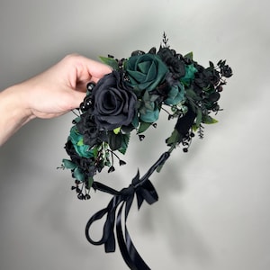 May include: A floral crown with dark green and black roses, complemented by small black flowers and green leaves. It is finished with a black satin ribbon. This gothic-style headpiece is suitable for special events.