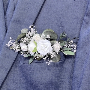 May include: A white flower boutonniere with greenery and small white flowers. The boutonniere is pinned to a dark blue suit jacket.