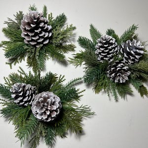 May include: Three decorative arrangements featuring natural brown pinecones with white-tipped edges, nestled among clusters of green artificial evergreen needles and cedar-like foliage, creating a textured, layered botanical display.