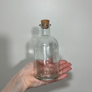 Op de afbeelding: Een heldere glazen fles met een kurken stopper. De fles wordt in een hand gehouden tegen een effen witte achtergrond. De fles heeft een afgeronde body en een smalle hals.