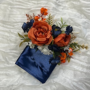 Puede incluir: Una tela de satén azul marino con un arreglo floral de rosas naranjas y blancas, flores azules y blancas y hojas verdes.