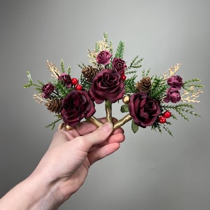 Puede incluir: Una corona floral con rosas burdeos oscuras, pequeñas piñas y bayas rojas. El arreglo incluye detalles dorados y follaje verde, creando un diseño festivo. La corona es sostenida por una mano.