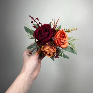 May include: A cluster of artificial flowers featuring a deep burgundy rose, an orange rose, and a smaller orange flower. Green leaves and sprigs of foliage surround the blooms. The arrangement is held against a light gray background.