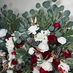 May include: A lush floral arrangement featuring white and red roses, green eucalyptus leaves, pine needles, and pine cones. Gold accents and red berries add festive touches. The arrangement is perfect for holiday decor.