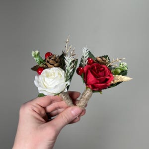 May include: Two small floral arrangements, one with a white rose and one with a red rose. Both arrangements include pine cones, berries, and greenery.