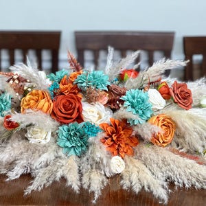 Op de afbeelding: Een bloemstuk met kunstbloemen en pampasgras. De opstelling omvat oranje, turquoise en witte bloemen, met een basis van pluizig, beige pampasgras. Het middelpunt staat op een houten tafel.