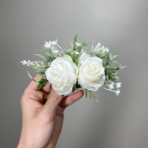 May include: A white floral arrangement with two large white roses and small white flowers, surrounded by green foliage.