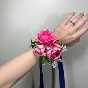 May include: A pink rose wrist corsage with a blue ribbon. The corsage is made of silk flowers and has a few white flowers and green leaves.