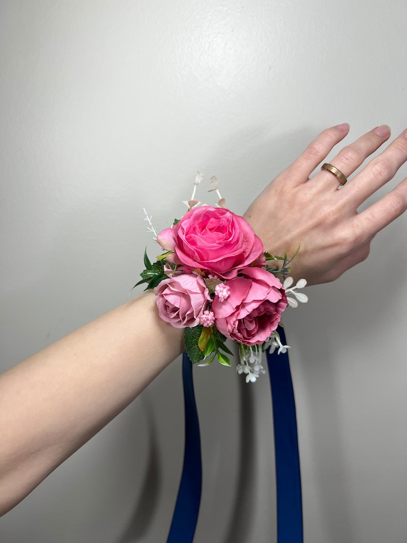 May include: A pink rose wrist corsage with a blue ribbon. The corsage is made of silk flowers and has a few white flowers and green leaves.