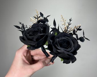 May include: Two black roses with gold accents and green leaves. The roses are arranged in a bouquet and are held in a hand.