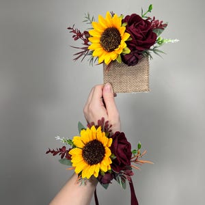 Puede incluir: Dos ramilletes de muñeca con un girasol amarillo y una rosa burdeos. Los arreglos florales están acentuados con vegetación y detalles burdeos, sujetos a una base de arpillera y cinta burdeos.
