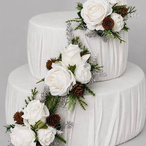 May include: A two-tier cake covered in white fabric with vertical pleats, decorated with clusters of white roses, small brown pinecones, green evergreen sprigs, and metallic silver foliage arranged in a diagonal pattern across the tiers.