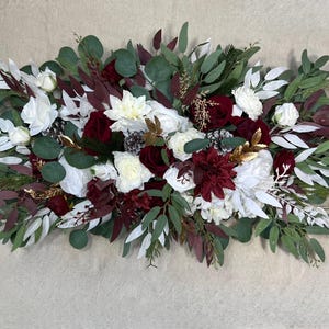 May include: A floral arrangement featuring white, burgundy, and cream-colored flowers, accented with green eucalyptus leaves and burgundy foliage. Gold accents and pinecones add detail to the centerpiece, creating a festive and elegant display.