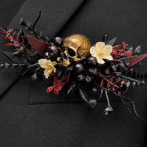May include: A decorative lapel pin featuring a central metallic gold skull surrounded by black foliage, dark red sprigs, and two small cream-colored flowers. The arrangement is composed of various artificial botanical elements and berries.