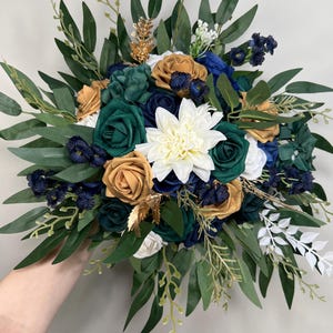 May include: A bouquet featuring a variety of artificial flowers in shades of green, navy, gold, and white. The arrangement includes roses, a large white dahlia, and greenery. The bouquet is held by a person.
