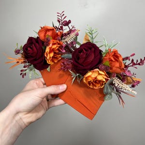 Puede incluir: Un arreglo floral con rosas burdeos, naranja y melocotón, follaje verde y tallos marrones. El arreglo está sobre un par de servilletas de tela naranja.