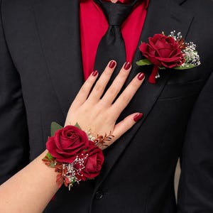 May include: A black suit with a red shirt and black tie. A red rose boutonniere is pinned to the lapel. A matching red rose wrist corsage is worn on the wrist. The nails are painted red.