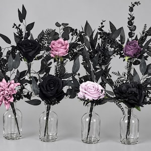 May include: A collection of four clear glass bottles, each containing a single artificial flower arrangement. The bouquets feature black foliage and stems paired with individual blooms in shades of black, dusty pink, and deep purple.