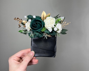 May include: A black satin square container filled with a floral arrangement. The arrangement includes a deep green rose, white roses, and small white flowers, with gold leaves and sprigs.