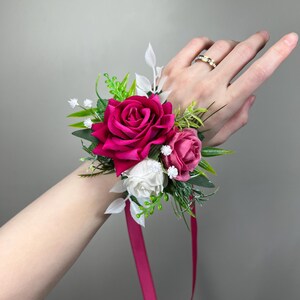 Puede incluir: Un ramillete de muñeca con una selección de flores vibrantes. El arreglo incluye una rosa grande de color rosa intenso, complementada con rosas rosas y blancas más pequeñas, follaje verde y acentos blancos. Una cinta rosa a juego asegura la pieza floral.