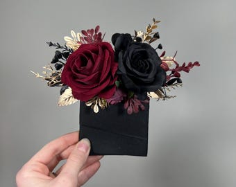 May include: A black and burgundy floral arrangement with gold accents. The flowers are arranged on a black ribbon.
