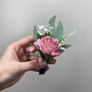 May include: A pink rose boutonniere with white and pink flowers and green leaves. The boutonniere is tied with a blue ribbon.
